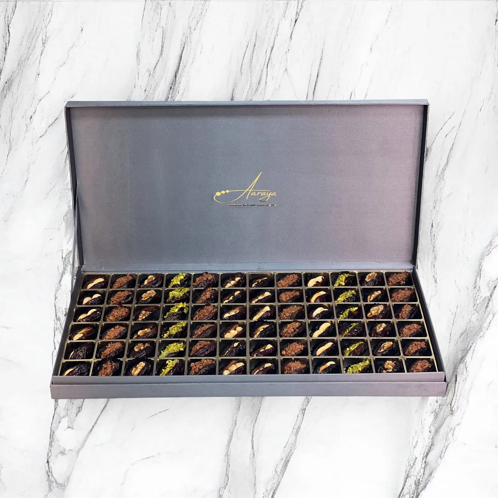 An exquisite assortment of Assorted Luxury Dates by Aaraya Chocolates, showcasing a symphony of premium flavors including Lemon Peel, Pistachio, Orange Peel, Cashew Nut, Salted Hazelnut, Feuilletine Crunch, Walnut, Almond Blanched, and Pistachio Ganache. Each date is a masterpiece of indulgence, promising a sensory journey through the finest gourmet fillings.