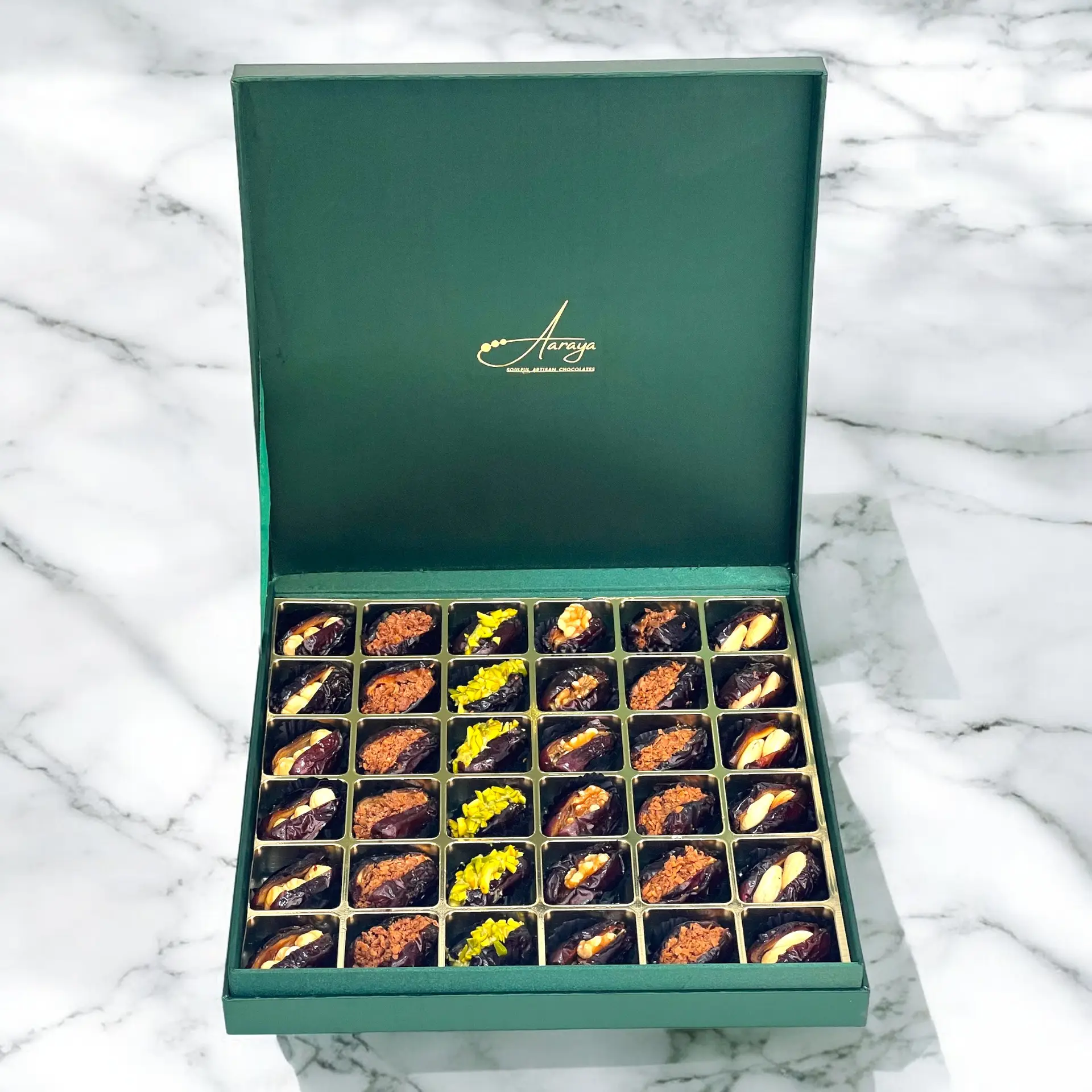 An exquisite assortment of Assorted Luxury Dates by Aaraya Chocolates, showcasing a symphony of premium flavors including Lemon Peel, Pistachio, Orange Peel, Cashew Nut, Salted Hazelnut, Feuilletine Crunch, Walnut, Almond Blanched, and Pistachio Ganache. Each date is a masterpiece of indulgence, promising a sensory journey through the finest gourmet fillings.